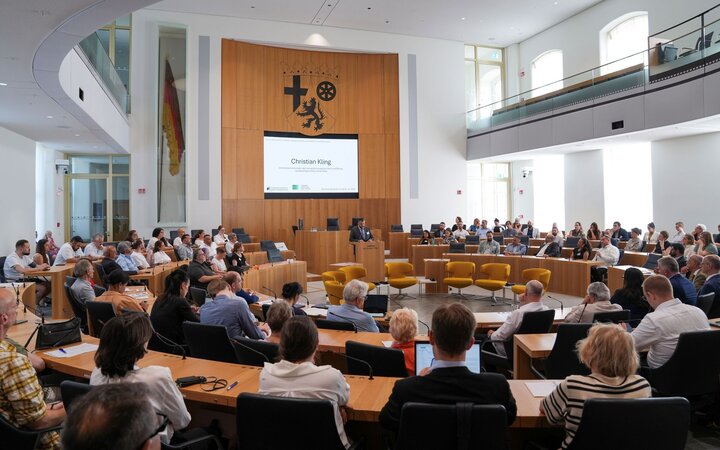 Foto vom Plenarsaal des Landtags Rheinland-Pfalz mit Kongressteilnehmenden und am Rednerpult Christian Kling vom Landesverband Rheinland-Pfalz des Verbands Deutscher Sinti und Roma