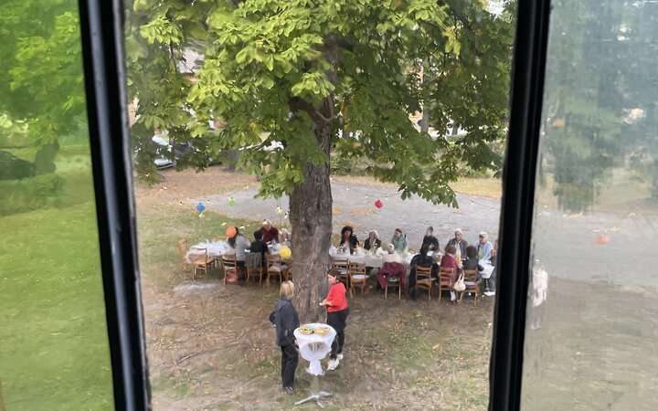 Blick von oben durch ein geteiltes Fenster mit Metallrahmen auf einen langen Tisch mit Menschen im Gespräch unter einem großen Laubbaum