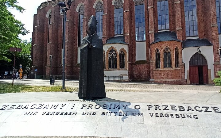 Statue eines Kardinals vor einer Backsteinkirche, davor am Boden die Worte "Wir vergebe und bitten um Vergebung"
