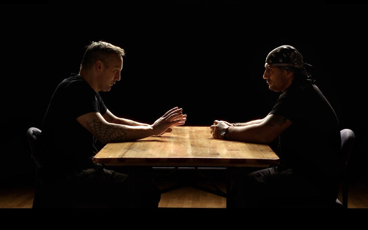 Zwei Männer in konzentrierter Haltung an einem erleuchteten Holztisch vor dunklem Hintergrund