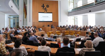 Foto vom Plenarsaal des Landtags Rheinland-Pfalz mit Kongressteilnehmenden und am Rednerpult Christian Kling vom Landesverband Rheinland-Pfalz des Verbands Deutscher Sinti und Roma