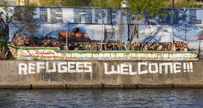 Betonmauer an einem Gewässer mit einem Graffito: Refugees Welcome