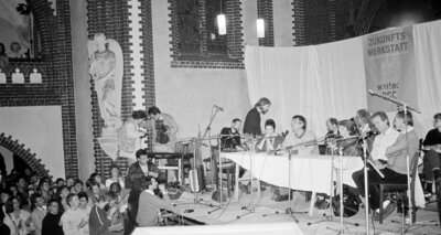 Podium mit improvisiertem Banner "Zukunftswerkstatt" im Altarraum einer Kirche, davor dicht gedrängte Menschen