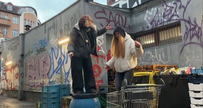 Zwei Jugendliche tanzen in einem Hinterhof mit Graffiti auf einer Tonne und einem Einkaufswagen